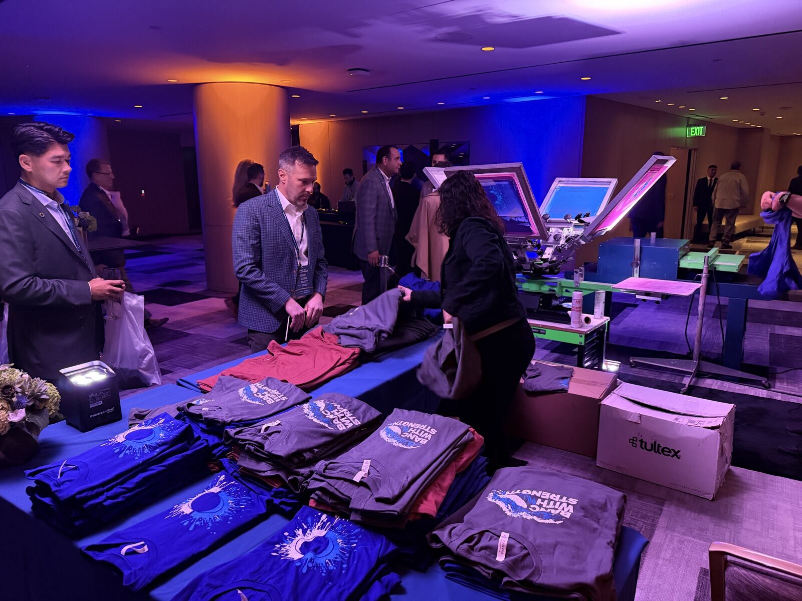 Corporate evening reception with folded navy and gray shirts stacked on a blue table beside the live station