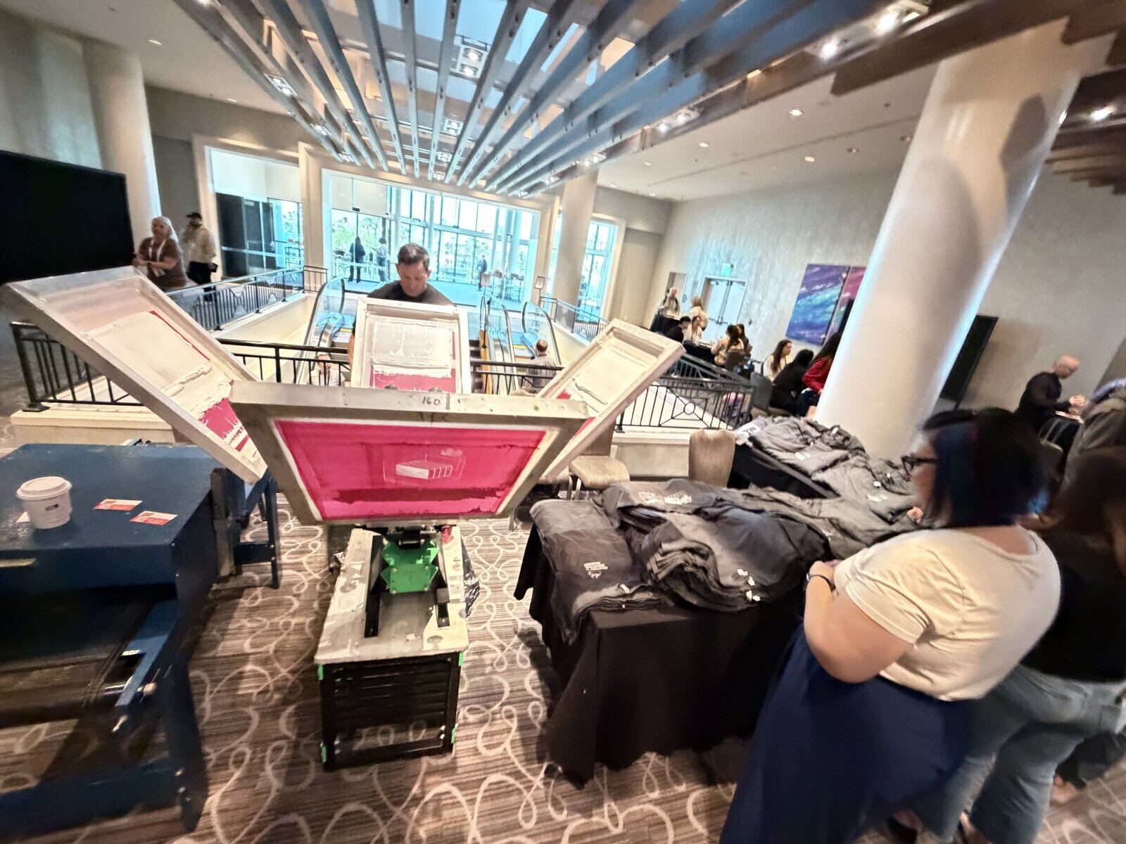 View over the equipment toward guests browsing a table stacked with folded gray shirts in a hotel lobby